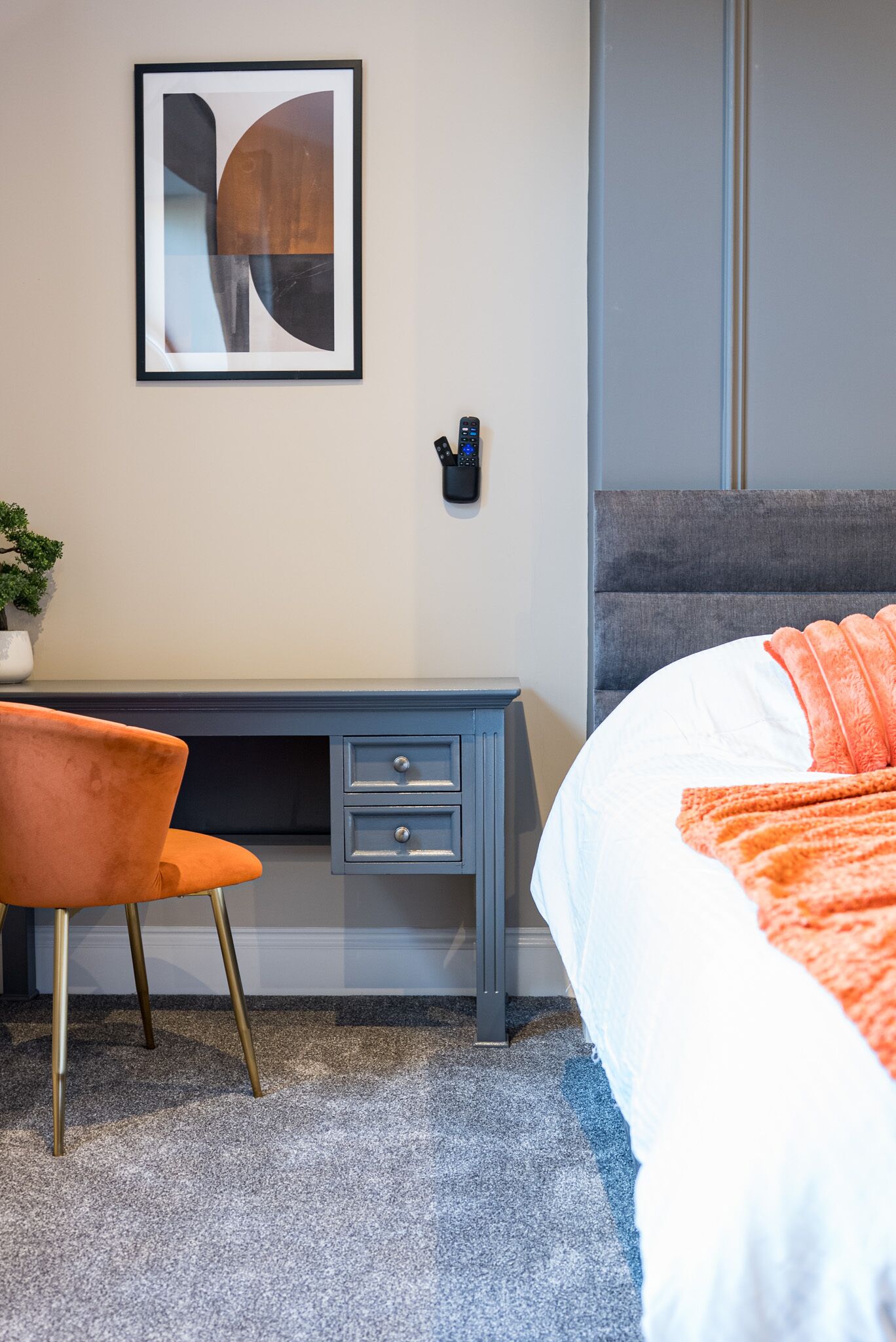 A hotel room with a bed, a desk with drawers, a chair, a framed abstract artwork, and a small potted plant.