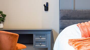 A hotel room with a bed, a desk with drawers, a chair, a framed abstract artwork, and a small potted plant.