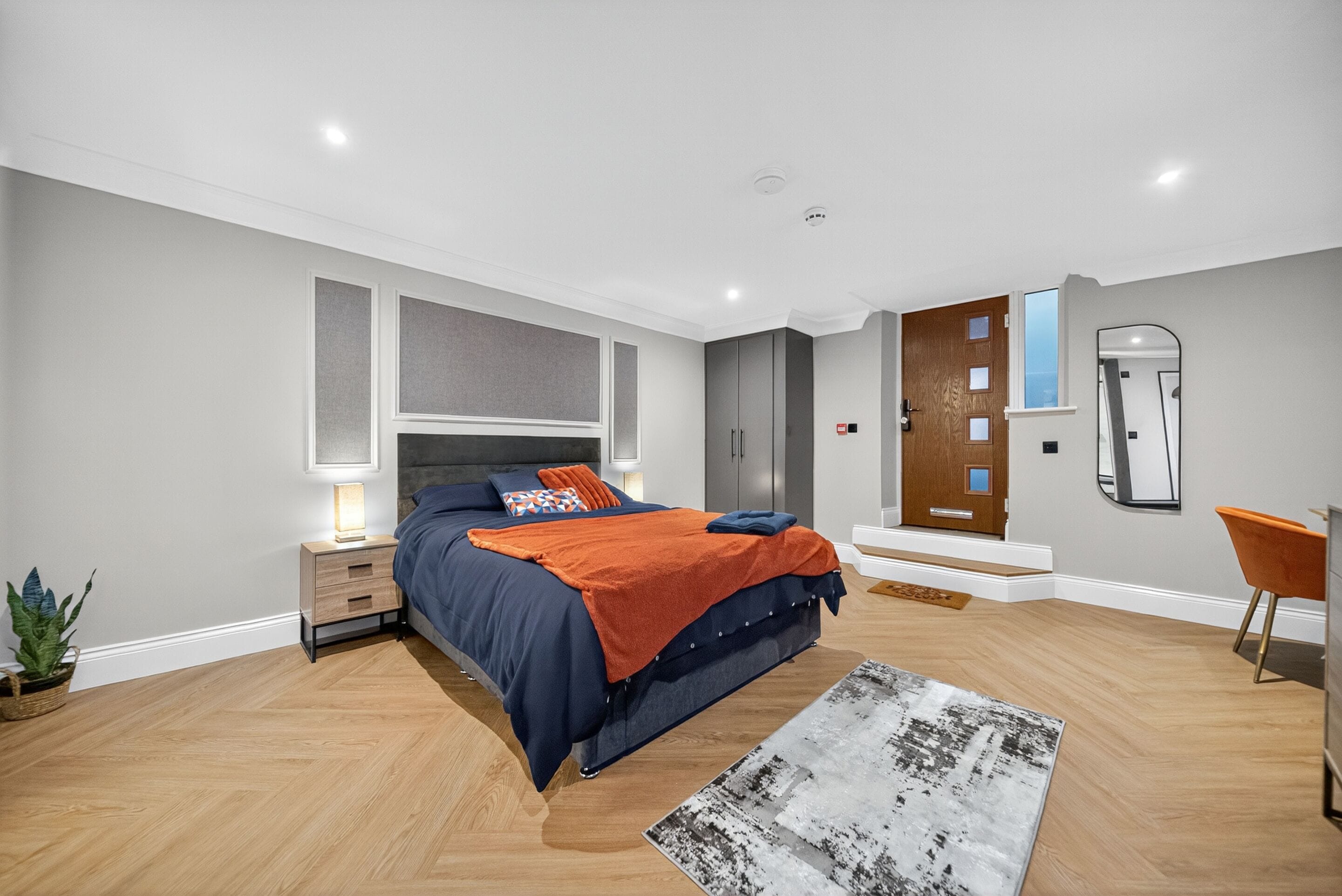 Un dormitorio con cama, mesita de noche, lámpara y un espejo en la pared.