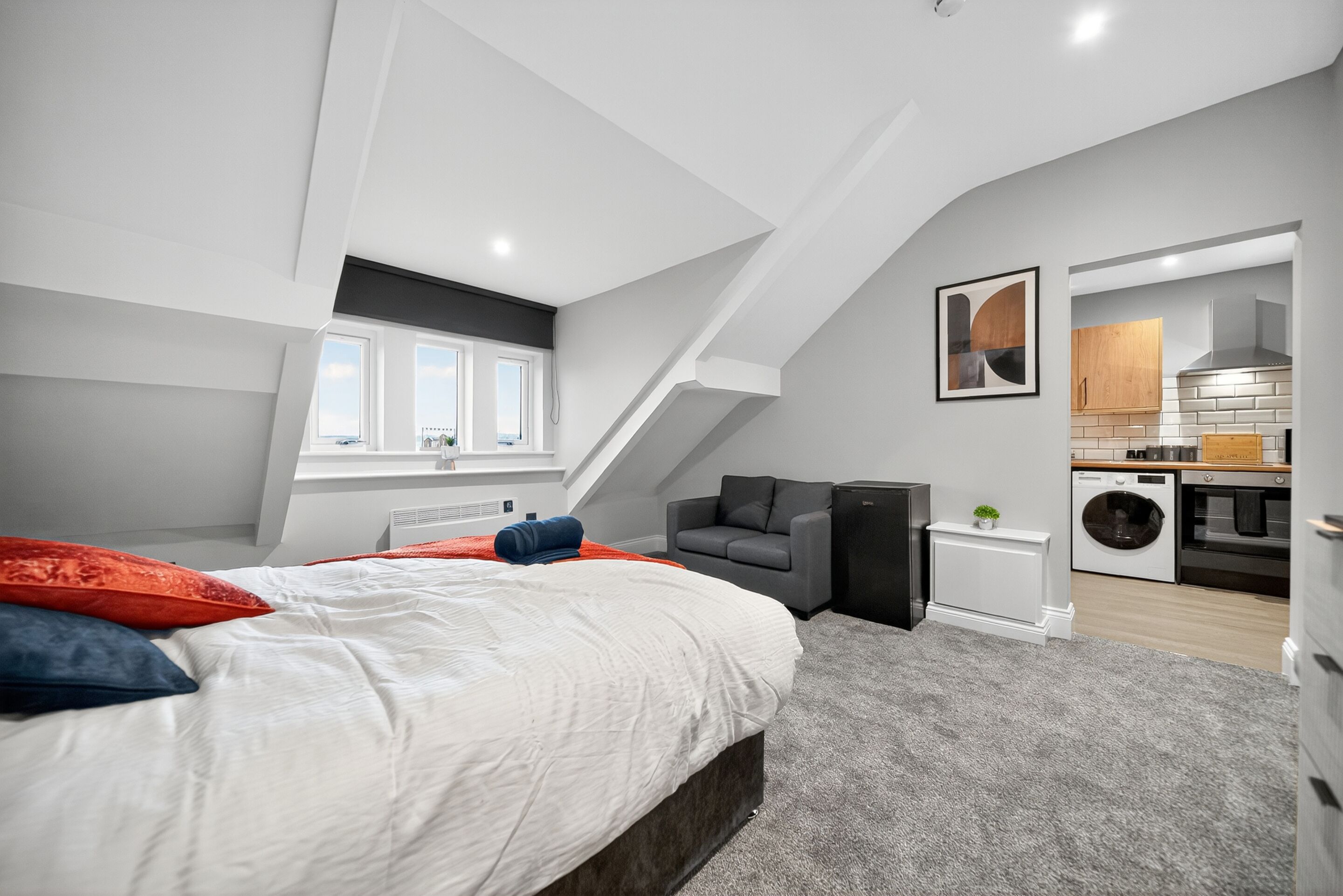 A modern bedroom with a sloped ceiling, a large bed with red and blue pillows, a gray armchair, and a view of the kitchen through an open doorway.