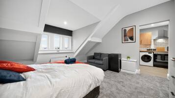 A modern bedroom with a sloped ceiling, a large bed with red and blue pillows, a gray armchair, and a view of the kitchen through an open doorway.