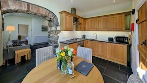 A modern kitchen with wooden cabinets, a stone archway, and a dining area with a table set for two. - Tan Y Bryn Y Beudy, Newport (Dinas Cross)