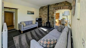 A cozy living room with a stone fireplace, a sofa, a television, and a dining area visible through an archway. - Tan Y Bryn Y Beudy, Newport (Dinas Cross)