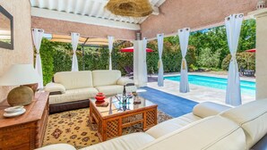 Ein überdachter Außenbereich mit Sitzgelegenheit, Pool, weißen Möbeln und einer hängenden Leuchte.