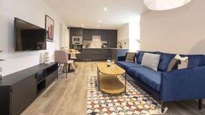 Una sala de estar moderna con un sofá azul, una mesa de centro, un televisor montado en la pared y una cocina al fondo.