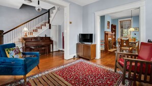 A spacious living room with a grand staircase, a piano, a television, and a dining area with chairs and a table. - Charming Historic Home-Walk to Carytown-Fence-Pet Friendly-Hotel-grade Towels (Richmond)
