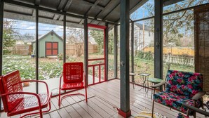 A covered porch with red chairs and a table, overlooking a backyard with a wooden fence and a green house. - Charming Historic Home-Walk to Carytown-Fence-Pet Friendly-Hotel-grade Towels (Richmond)