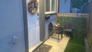 En bostadsfasad med en dörr, ett fönster, en liten uteplats med två stolar, en krukväxt och en häck.