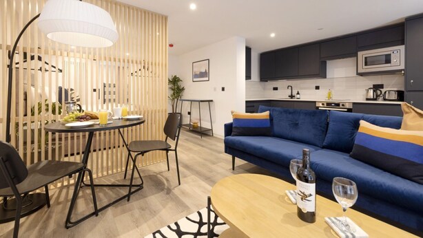 A modern living room with a blue sofa, a dining table with chairs, a kitchen area with dark cabinets, and a large window. - Argyl | Abbey Gate: Studio 3 (Edinburgh)