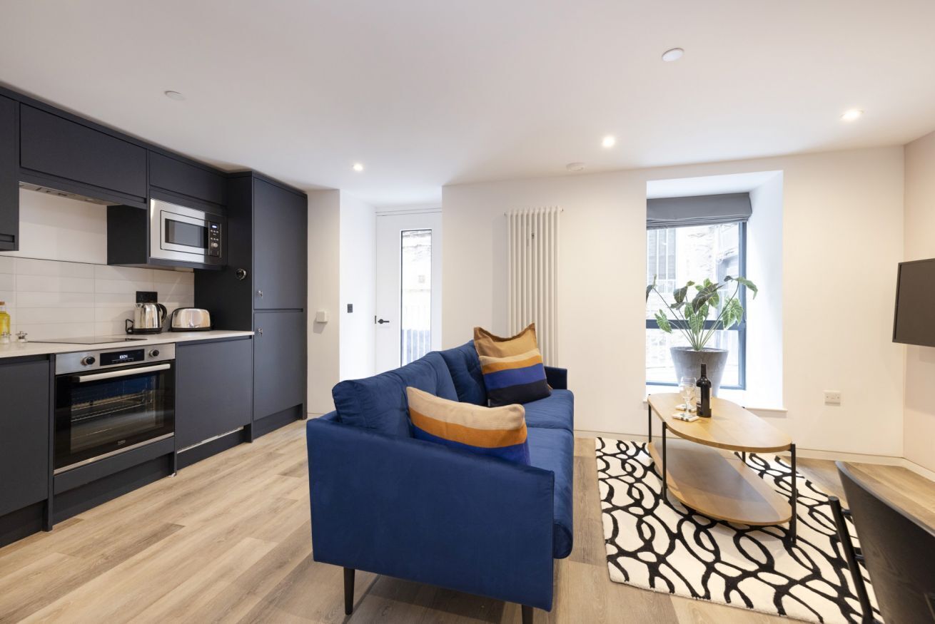A modern kitchen with dark cabinets, a microwave, and an oven. There's a blue sofa with cushions and a small table with a plant.