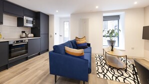 A modern kitchen with dark cabinets, a microwave, and an oven. There's a blue sofa with cushions and a small table with a plant. - Argyl | Abbey Gate: Studio 3 (Edinburgh)