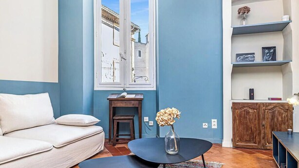 Una sala de estar con pared azul, un sofá blanco, una mesita de madera, un jarrón con flores y vista a la calle a través de una ventana.