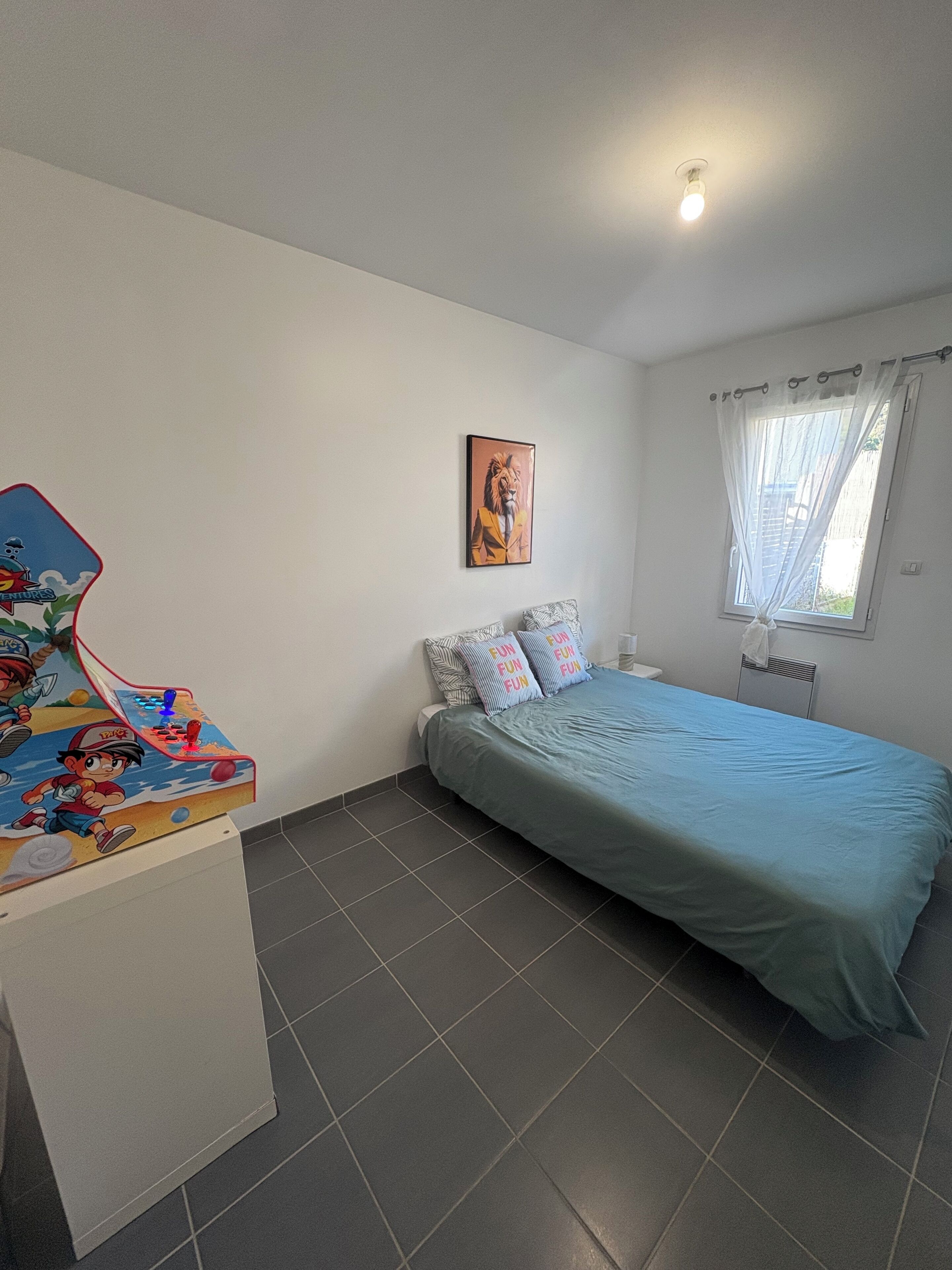A bedroom with a bed, a wall-mounted picture, a window with curtains, and a small desk with a cartoon character.