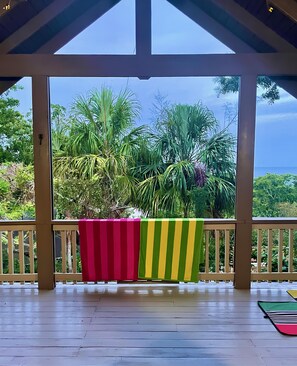 A covered outdoor area with a wooden floor and a view of lush greenery and the ocean. - Private 3-bedroom house a three minute walk to Luna Beach (Roatan)