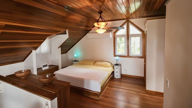 Ein Schlafzimmer mit schrägverdachter Decke, Holzboden, einem Bett, einem Nachttisch, einer Lampe und einem Fenster.