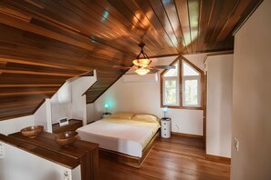 Un dormitorio con techo inclinado, piso de madera, cama, mesita de noche, lámpara y ventana.