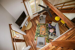 Una habitación con techo de madera, ventilador de techo, ventana con cortinas, televisor, una pequeña mesa con un juego de mesa y un sofá con cojines estampados.
