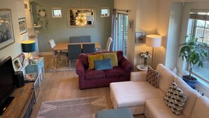 Ein helles Wohnzimmer mit einer Couch, Sesseln, einem Esstisch und einem Fernseher.