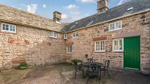 A stone building with a green door, a courtyard with a table and chairs, and a small garden area. - Beautiful Lake District Cottage With Log Burner (Penrith)