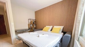 A neatly made bed with white linens and two pillows, a wooden slat wall behind it, and a small side table with a lamp.