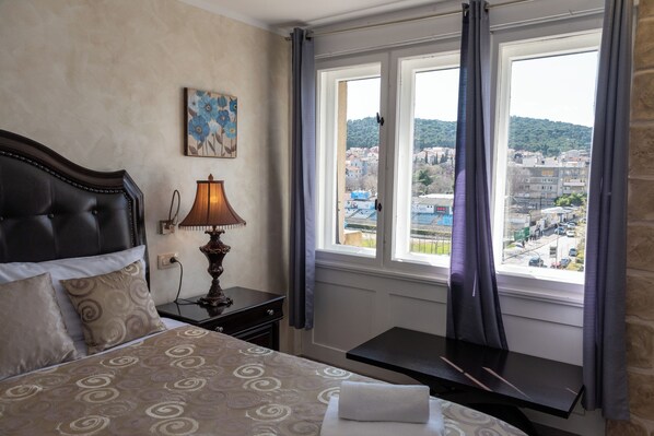 A hotel room with a bed, a nightstand with a lamp, a small table, and a window with curtains. - Queen Mary Luxury Rooms Split (Split)