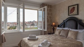 A bedroom with a bed, a nightstand, a lamp, a window with curtains, and a city view. - Queen Mary Luxury Rooms Split (Split)