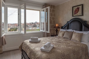 Ein Schlafzimmer mit Bett, Nachttisch, Lampe, einem Fenster mit Vorhängen und Blick auf die Stadt.