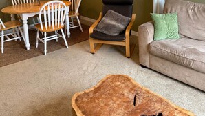 Una sala de estar con una mesa de centro redonda de madera, un sofá color beige con un cojín verde, una zona de comedor con una mesa y sillas blancas, y un cuadro colgado en la pared que representa un ciervo.