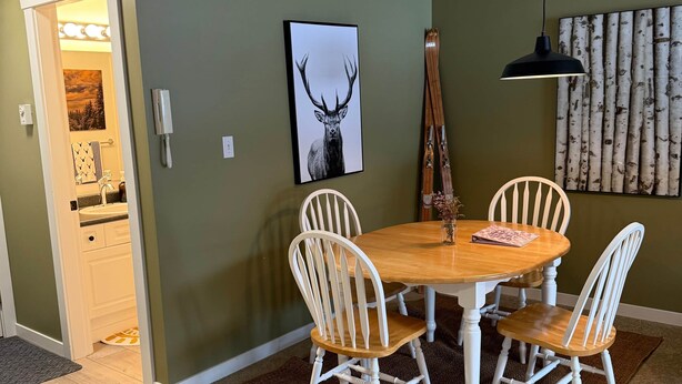 Un comedor con una mesa de madera y sillas blancas, un cuadro de un ciervo en la pared, y un adorno mural de madera de abedul.