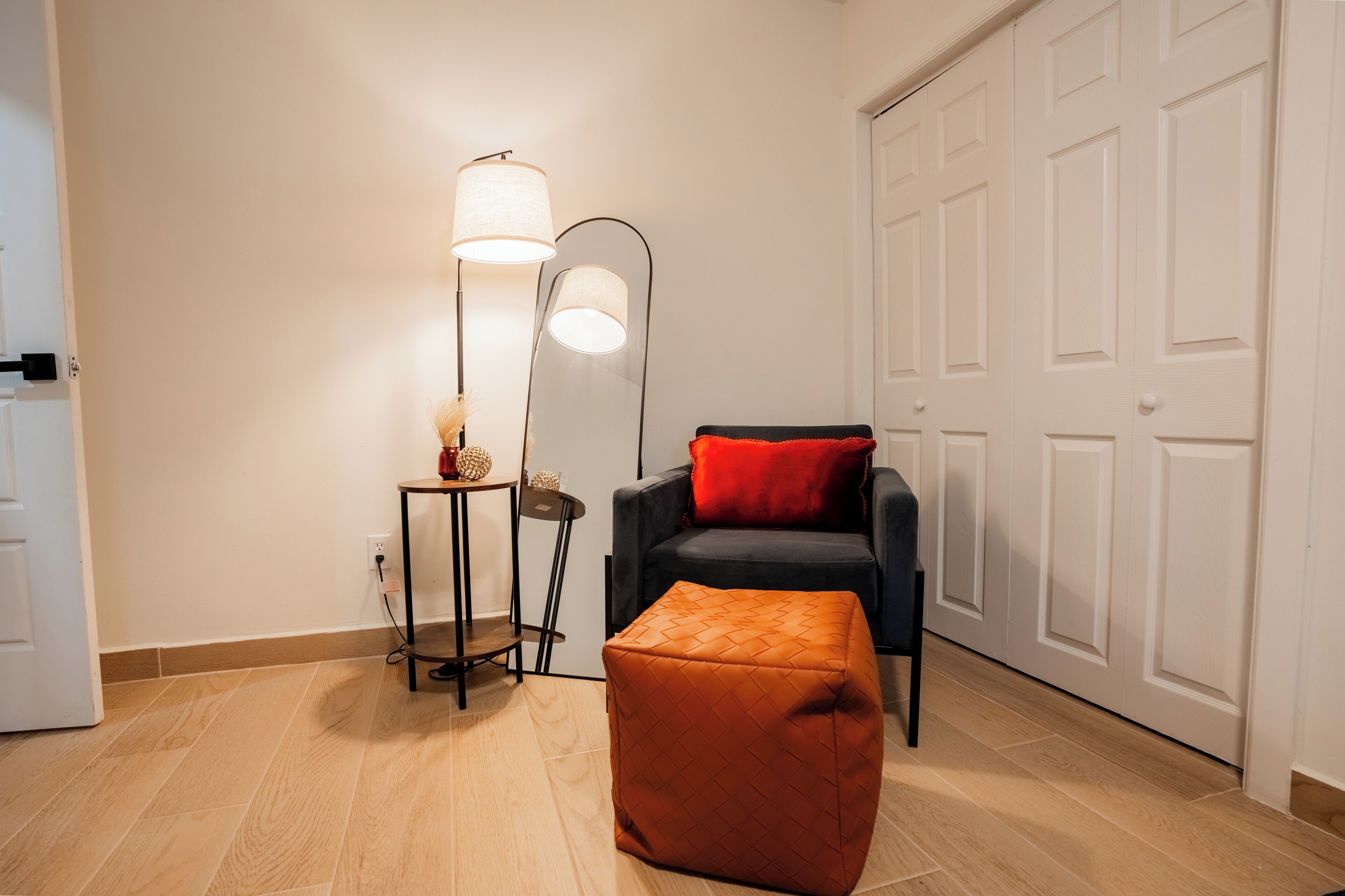 Una habitación con piso de madera, una silla negra con cojín rojo, una lámpara de pie alta y una mesita con un florero.