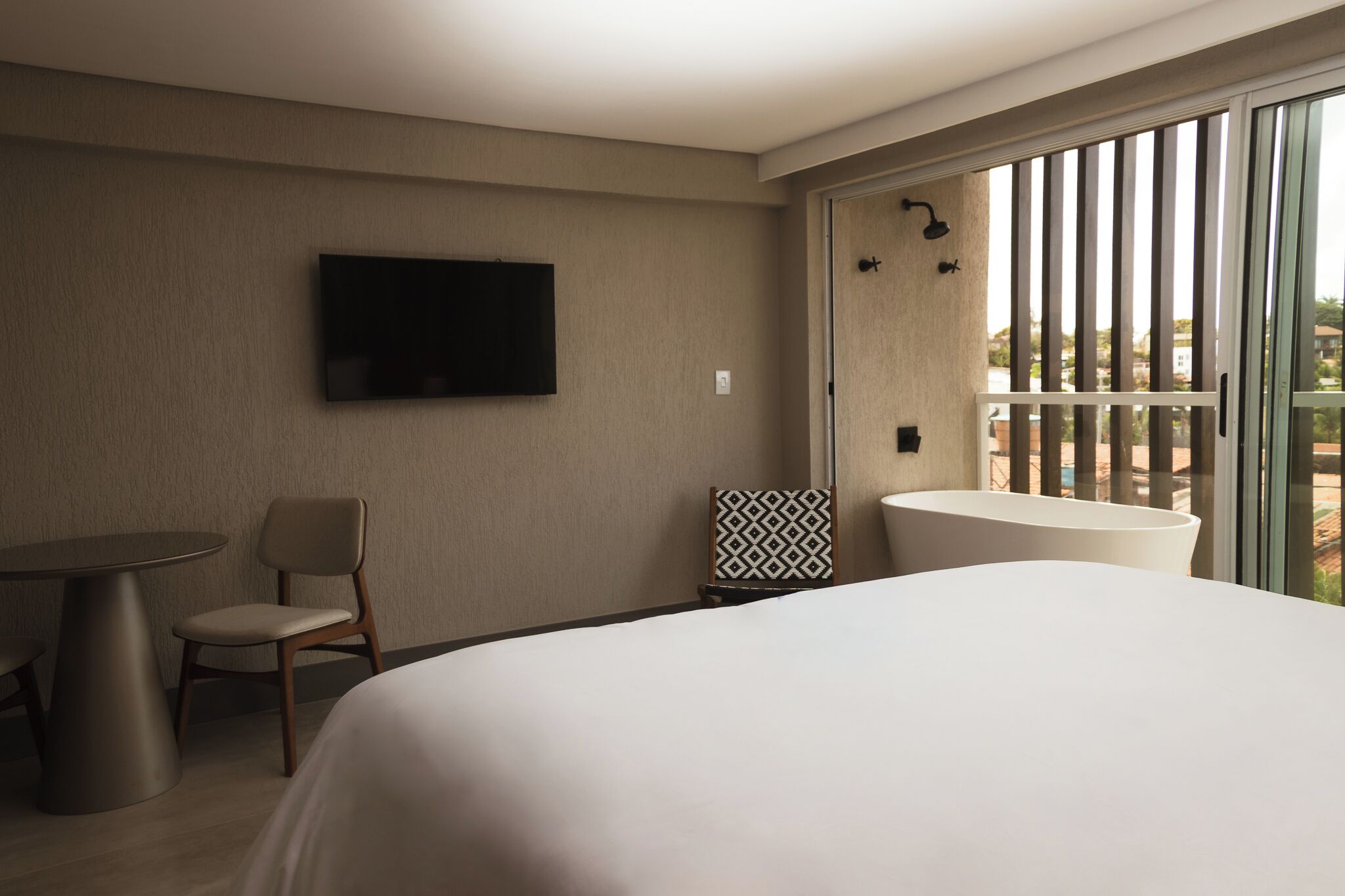 A hotel room with a bed, a TV mounted on the wall, a chair, a small table, a patterned pillow, and a balcony with a view.