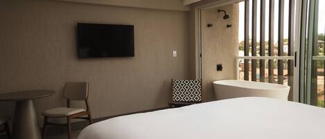 A hotel room with a bed, a TV mounted on the wall, a chair, a small table, a patterned pillow, and a balcony with a view.