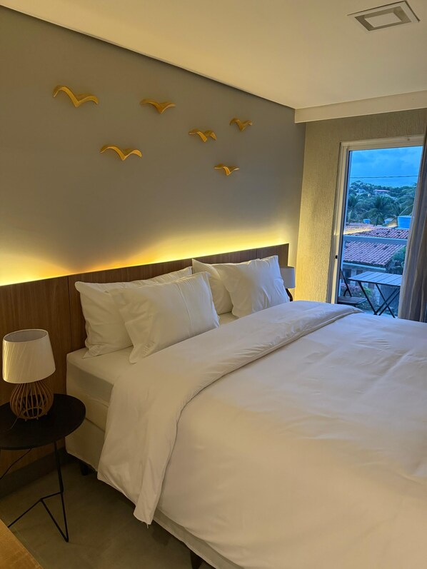 Habitación de hotel con una cama grande, una mesita de noche con lámpara y vistas al exterior.