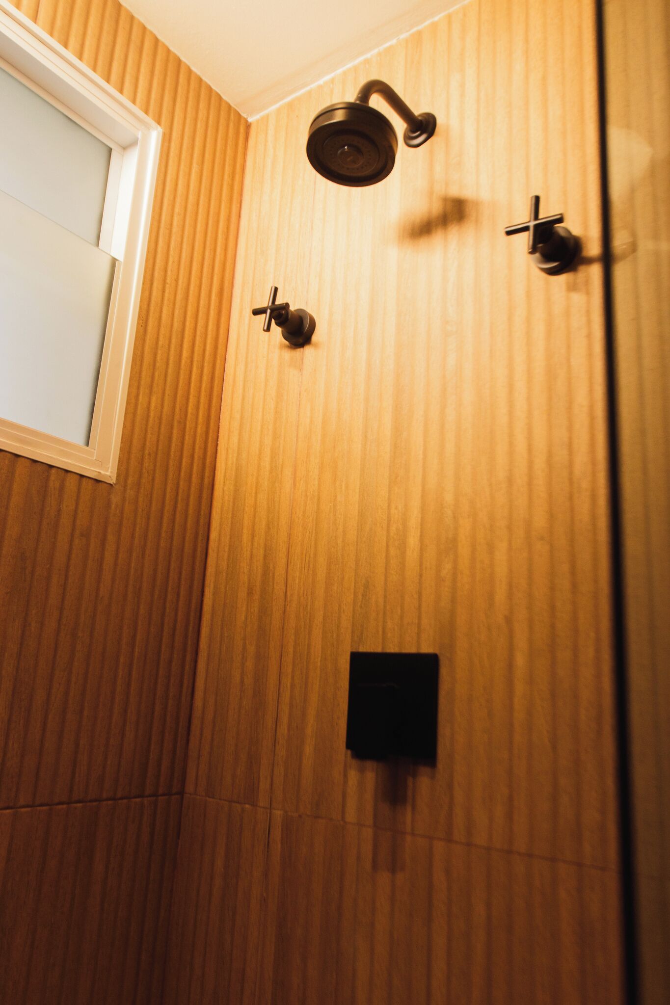 Ducha con pared de paneles de madera, un pequeño panel de control negro y una ventana con marco blanco.