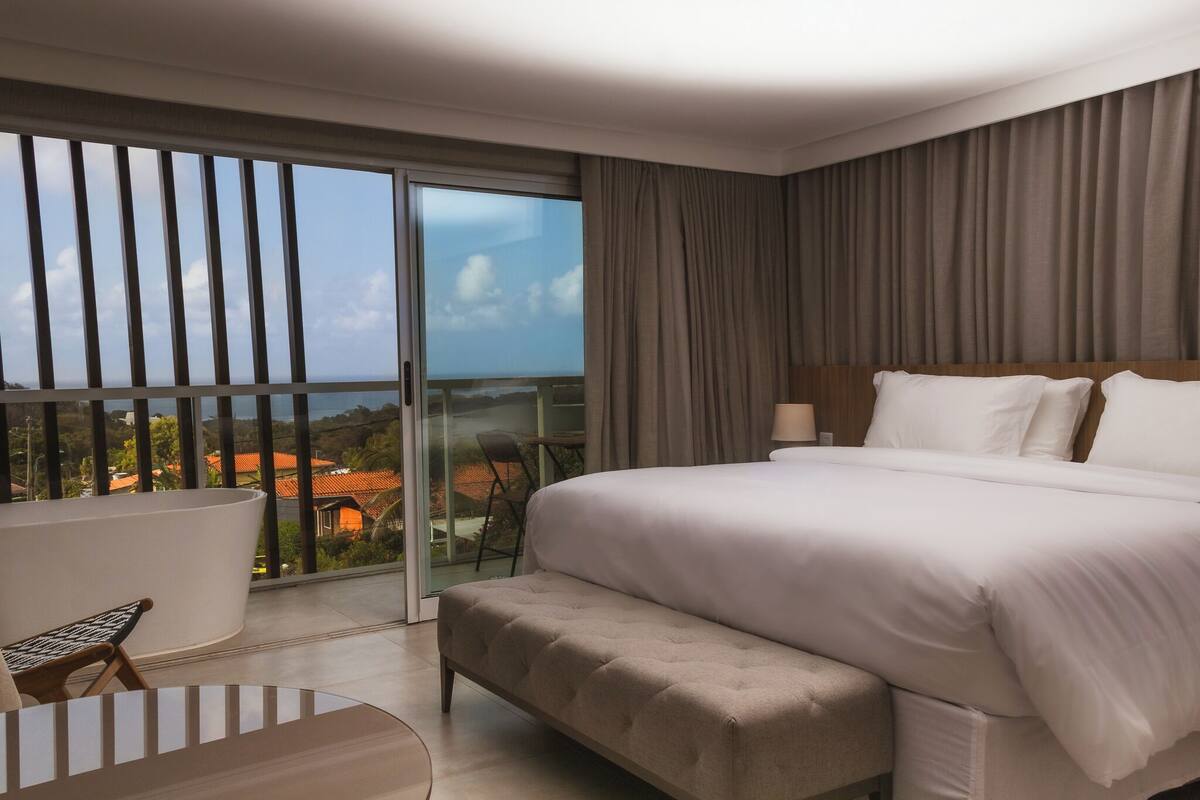 A modern hotel room with a large bed, a sitting area with a chair, and a balcony with a view of the city.