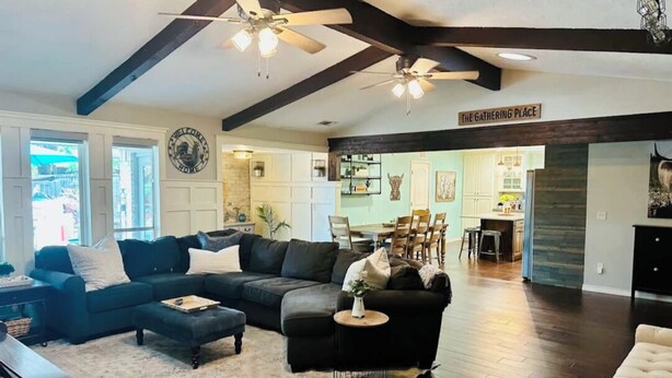 A spacious living room with a sectional sofa, a round coffee table, and a piano. There is a dining area with a table and chairs visible in the background.