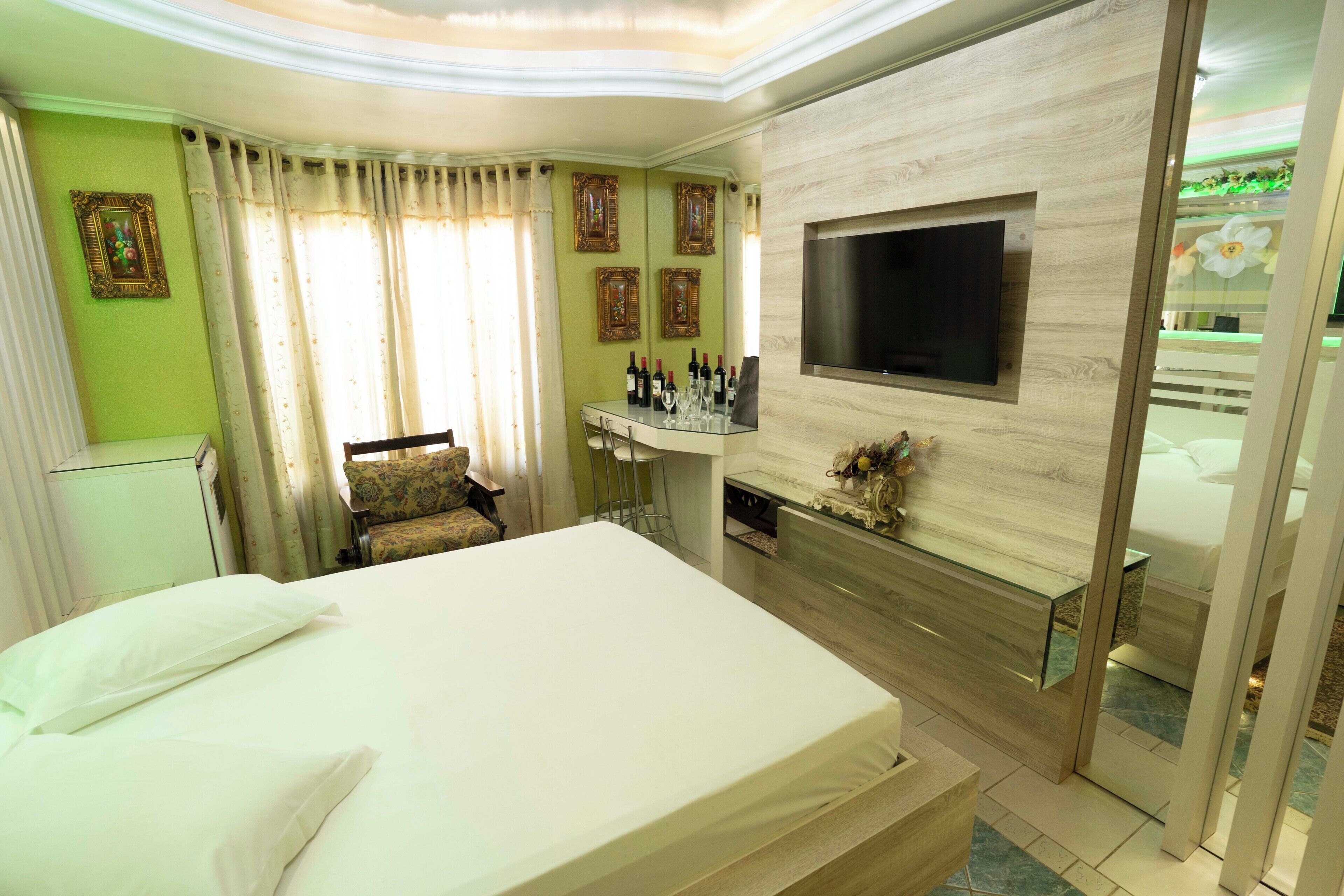 A hotel room with a bed, a TV mounted on the wall, a small table with bottles, and a chair.