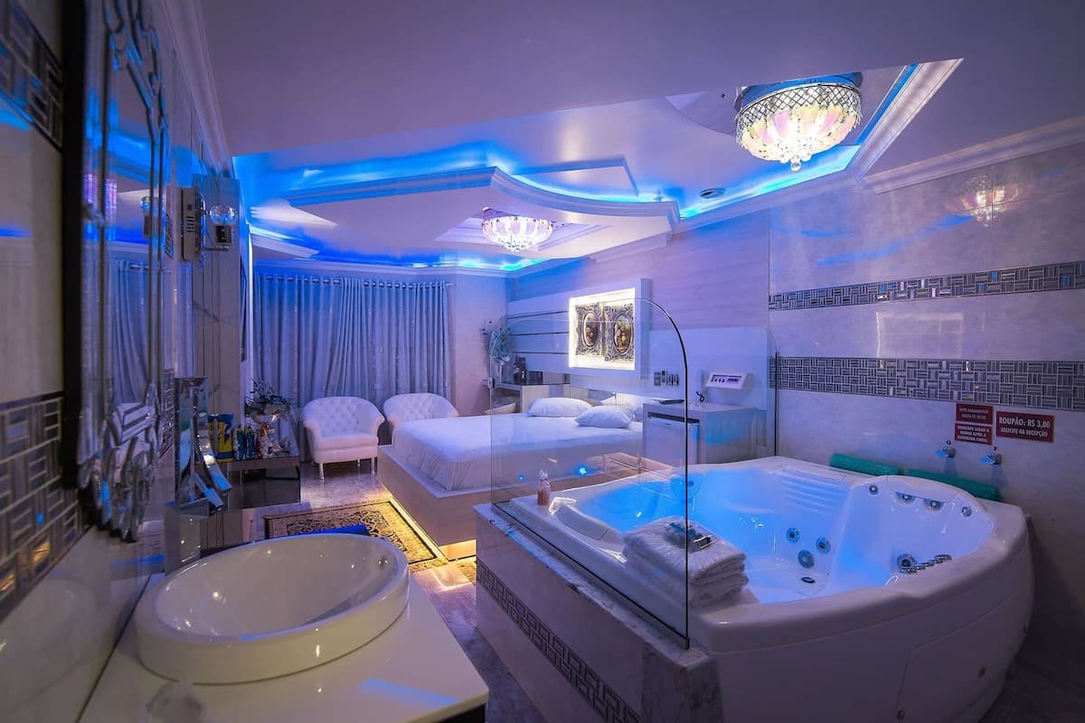 A modern hotel room with a large bathtub, a separate bedroom with a bed, a sitting area with chairs, and a bathroom with a sink and mirror.