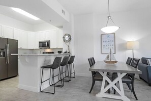 A modern kitchen with white cabinets, a stainless steel refrigerator, a microwave, and a dining area with a table and chairs. - The Palms Destin 2103 (Destin)
