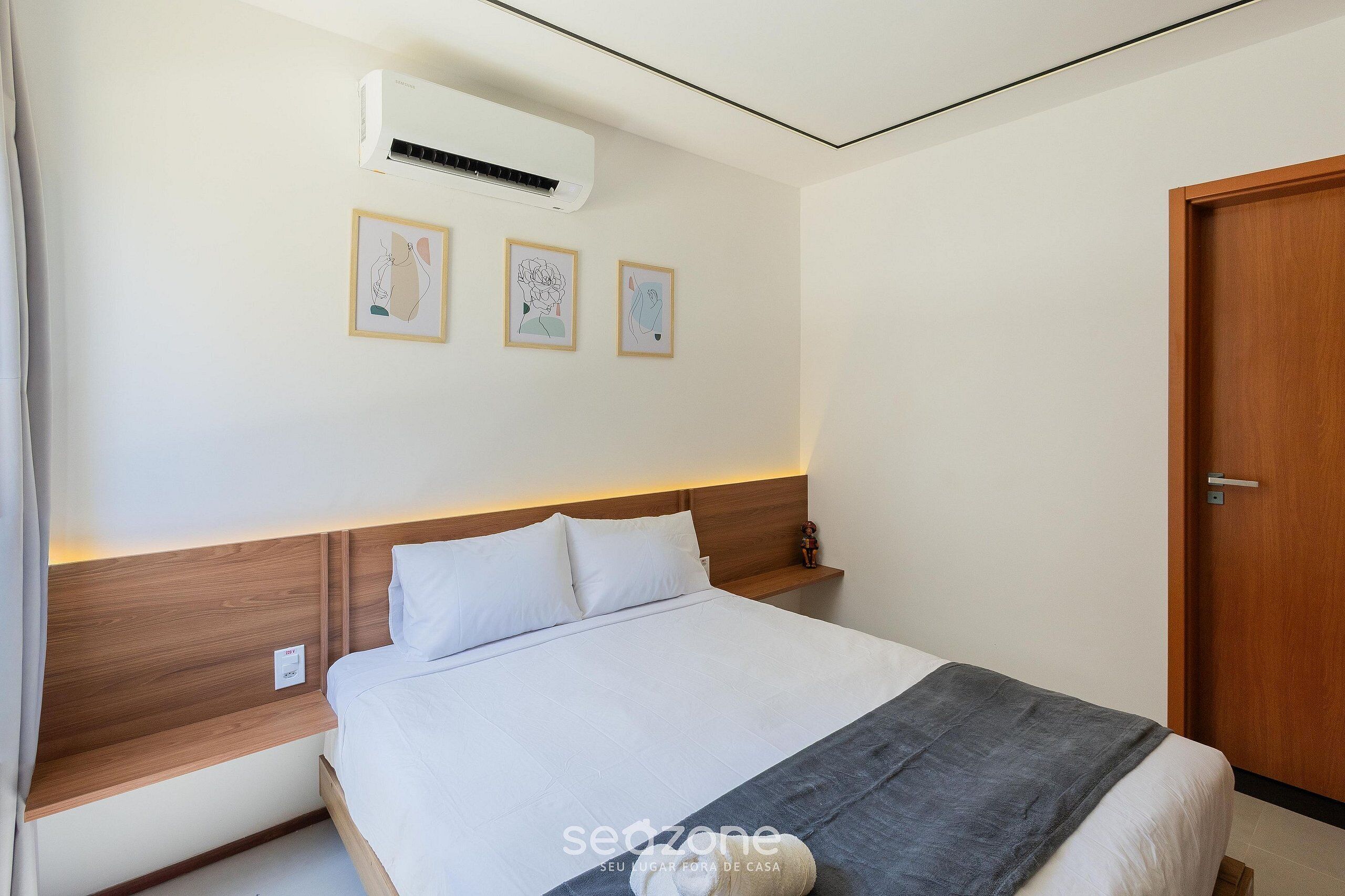 A neatly made bed with white linens, a grey throw blanket, and a wooden headboard. There are three framed artworks on the wall above the bed.