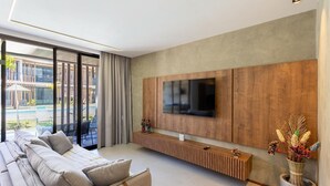 A modern living room with a large TV mounted on a wooden panel wall, a sofa with cushions, a console table, and a view of an outdoor pool area. - Apartments in Condo With Pool in Front of the Beach - NVB (Porto de Pedras)