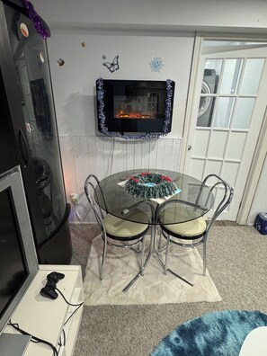 Un comedor con una mesa de vidrio rodeada de sillas, un televisor montado en la pared y un refrigerador a la izquierda.