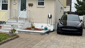 Una casa residencial con un coche negro estacionado en la entrada, un pequeño jardín con plantas en macetas y un buzón en la pared.