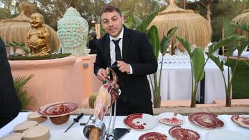 Een man in pak bereidt eten buiten, bij een gedekte tafel, omringd door decoratieve objecten.