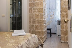 A hotel room with a stone accent wall, a shower with a glass door, a bed with a patterned bedspread, and a small seating area with a chair. - Queen Teuta Luxury Rooms Split (Split)