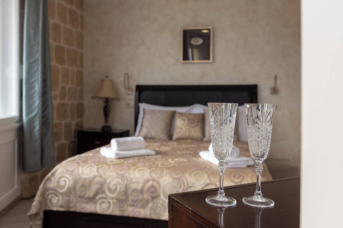 A hotel room with a bed, two bedside tables, a lamp, and two crystal glasses on a table.