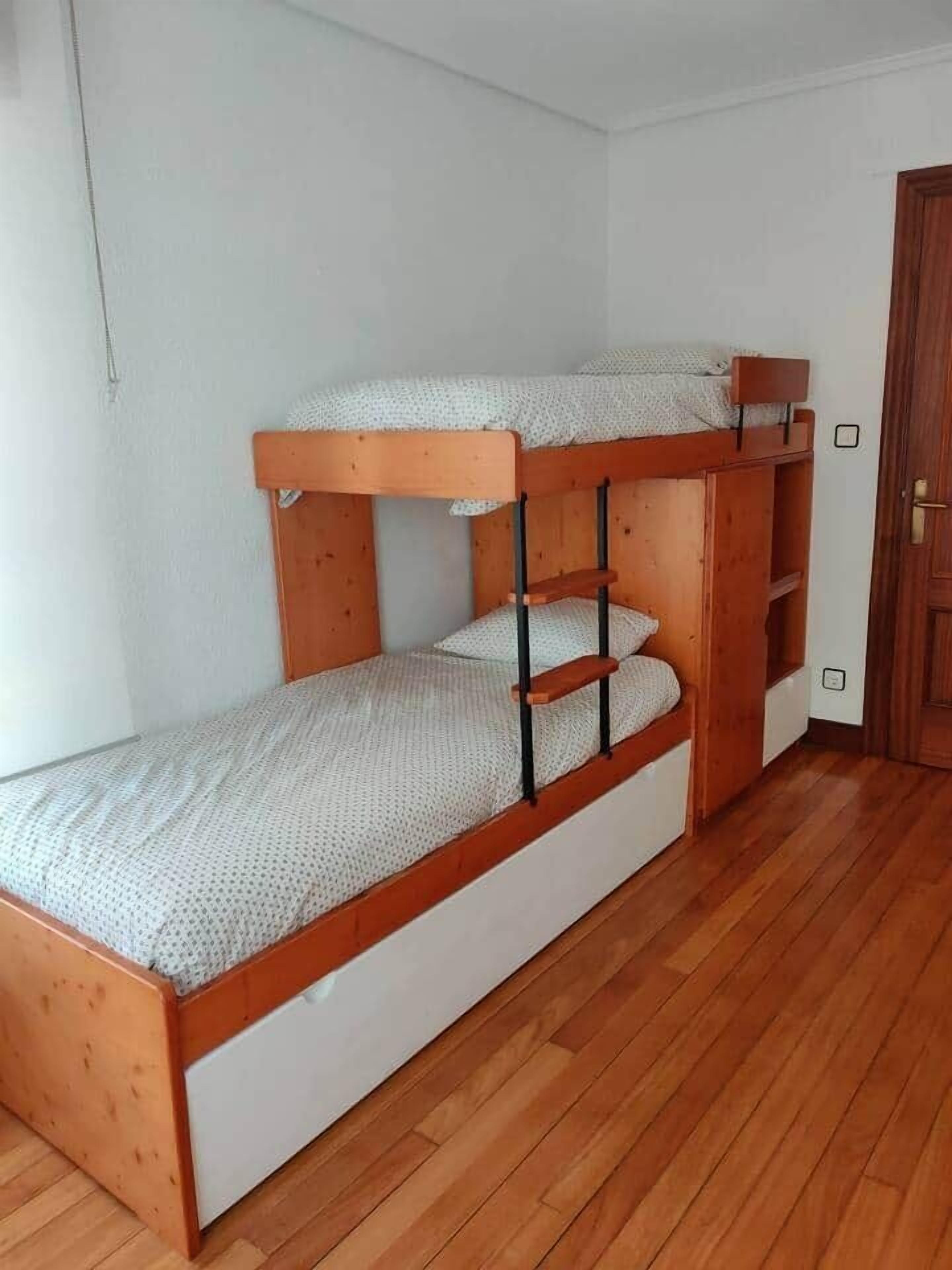 A bunk bed with a wooden frame and white bedding in a room with wooden flooring and a white wall.