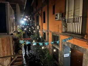 Un estrecho callejón flanqueado por edificios, algunos con balcones y unidades de aire acondicionado. Hay luces colgantes y un banderín azul cruzando el callejón.