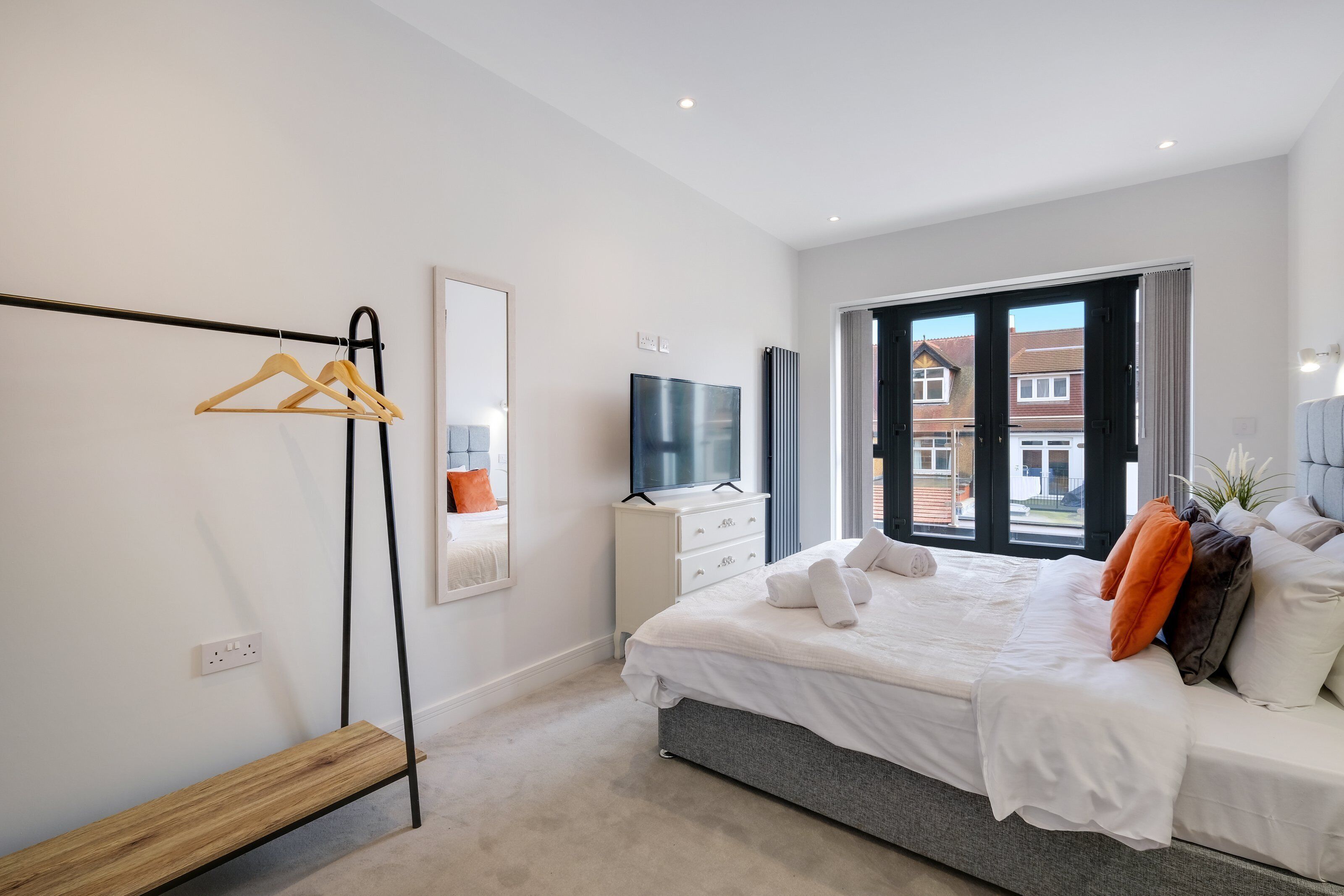 A modern bedroom with a bed, a TV, a mirror, and a window with a view of houses.
