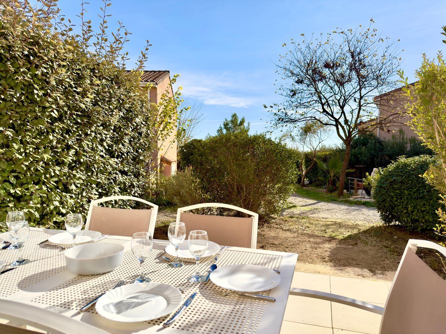 An outdoor dining area with white tables and chairs, surrounded by greenery and a gravel path.